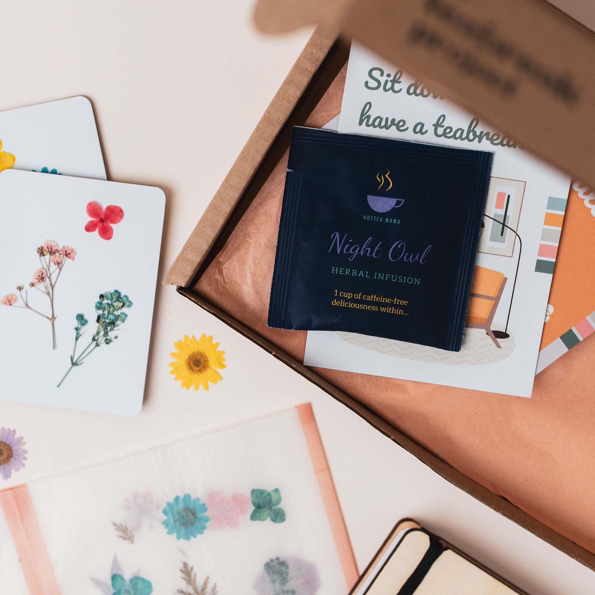 An opened Teabreak box showing a tea bag, illustrated cards and pressed flowers on a calm surface.