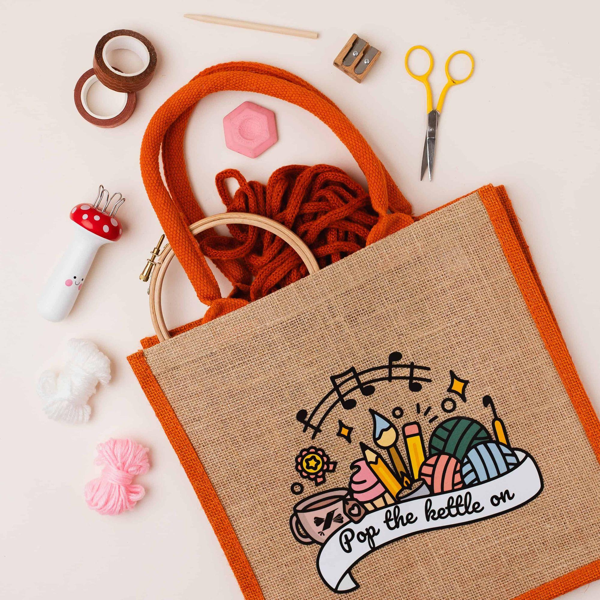 The Teabreak bag with Pop the kettle on printed on it, surrounded by an embroidery hoop, orange yarn, scissors, washi tape and colourful craft supplies